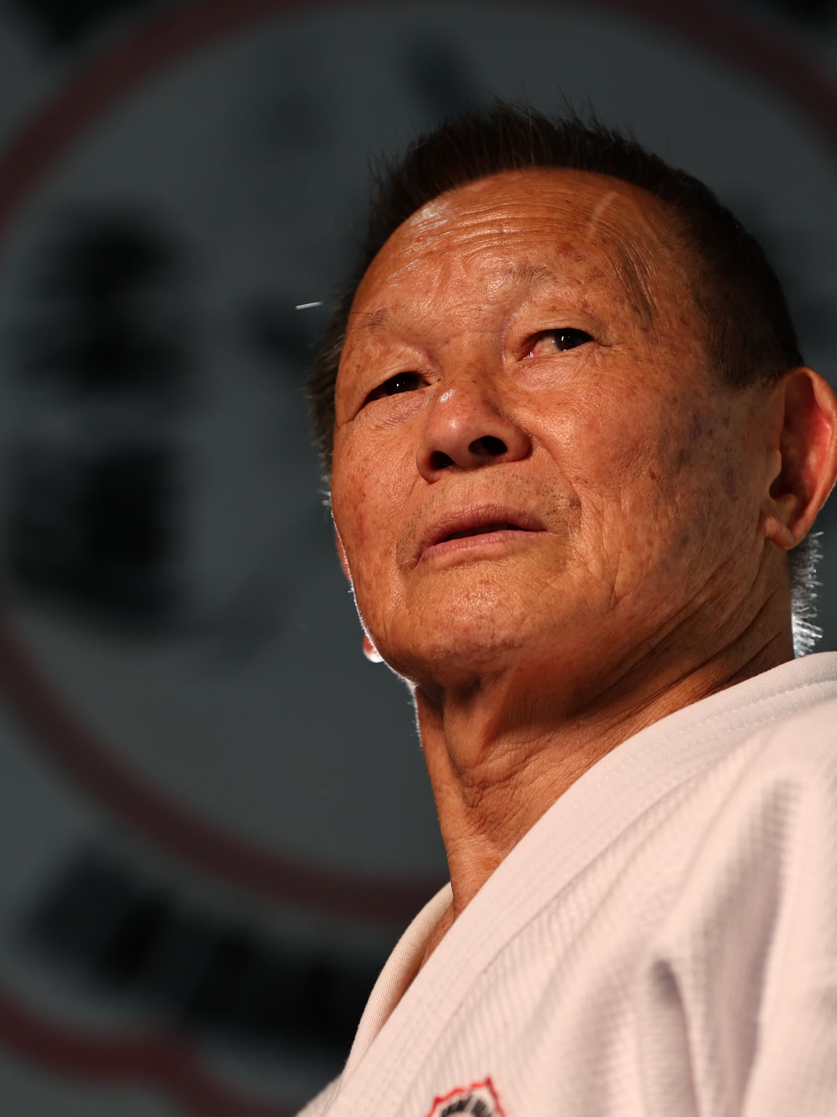 Sensei Sérgio Honda — olhar firme, kimono branco, crista do dojo ao fundo